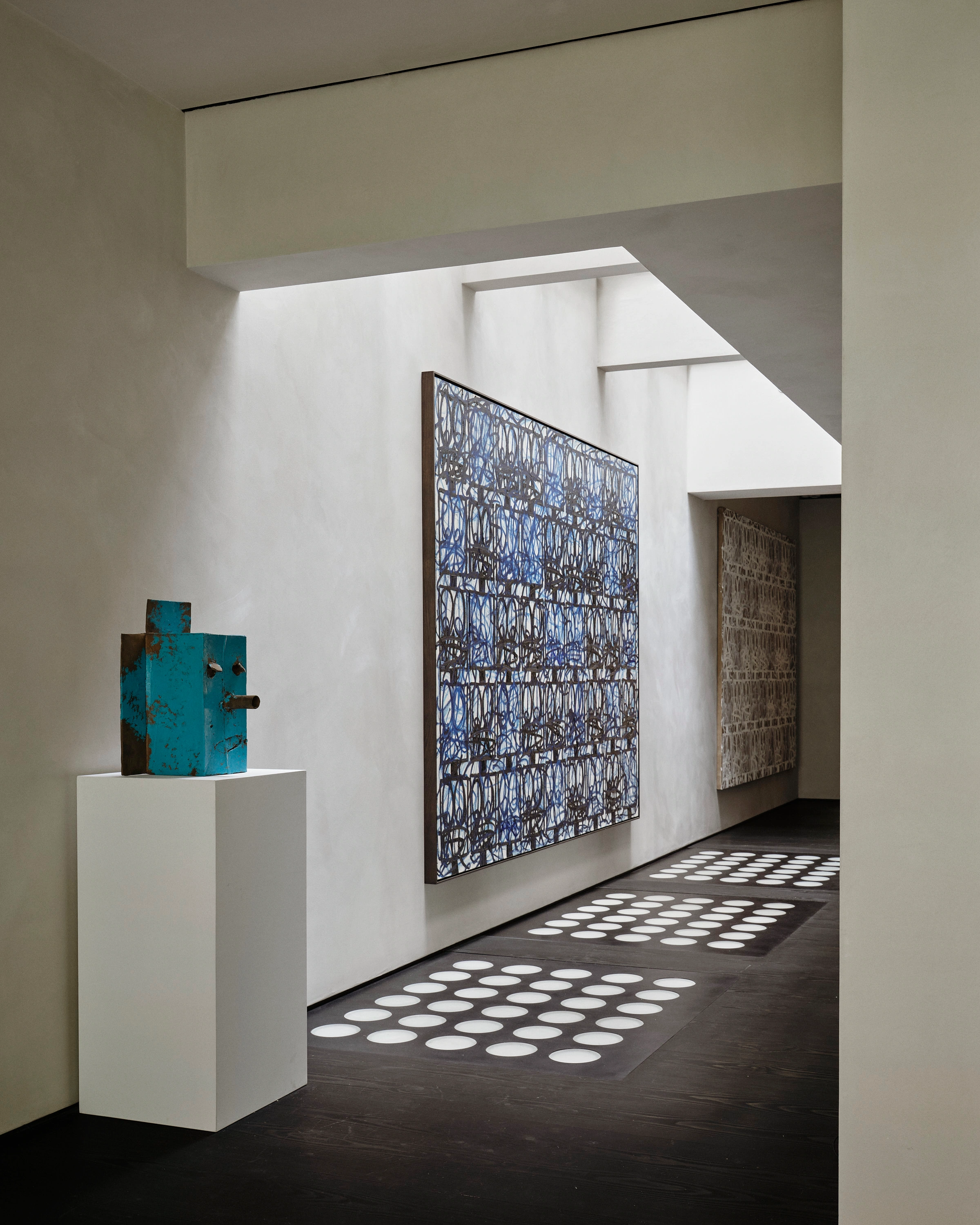 Gallery hallway with plaster walls, dark wood flooring dappled with circular light patterns, a small blue-painted sculptural totem on a white pedestal, and a large abstract painting with blue and gray linework hanging on the wall. Nicolas Schuybroek
