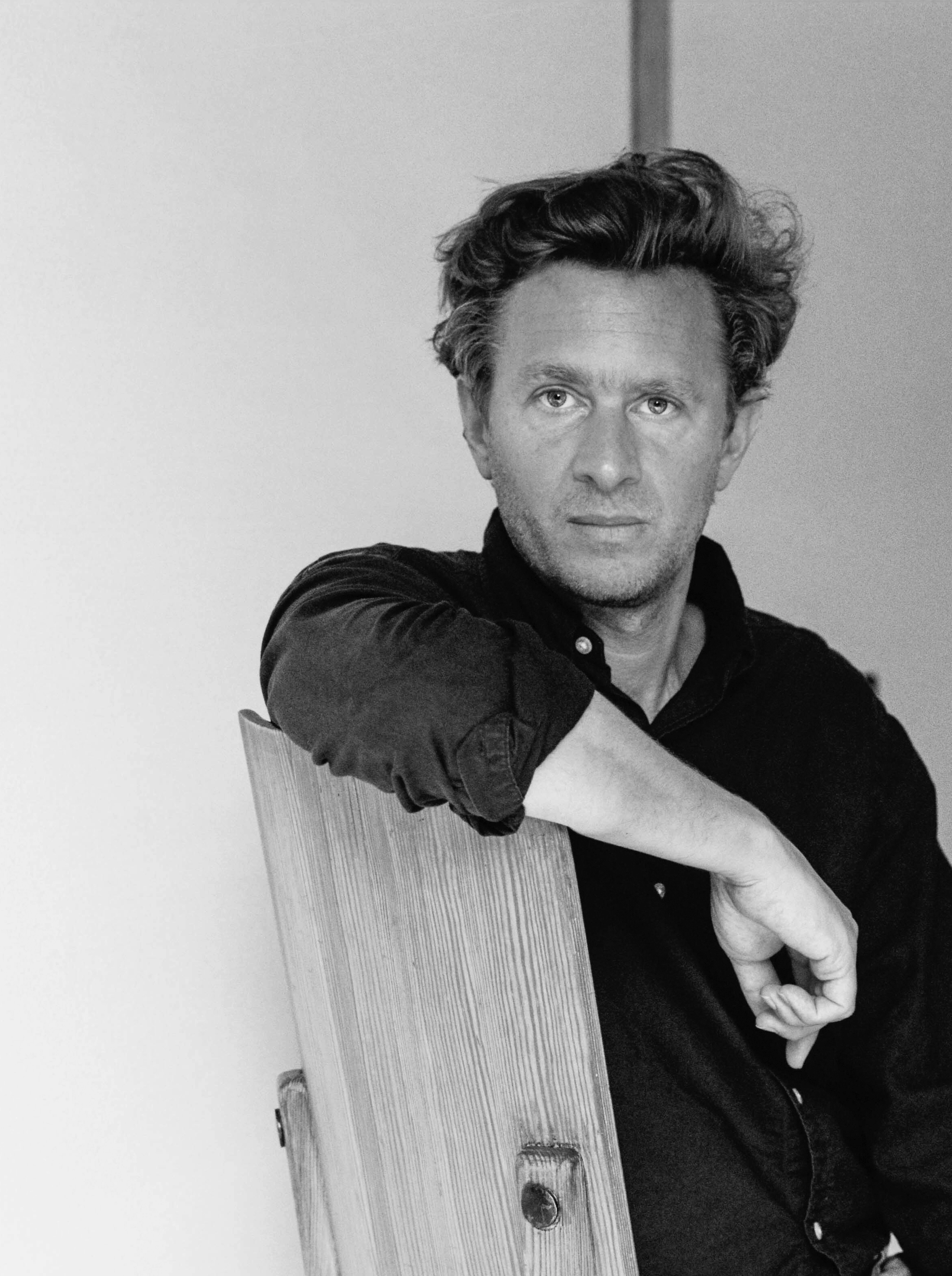 Black-and-white portrait of a man with wavy dark hair, wearing a dark shirt, leaning his arm on a tall wooden panel while gazing directly at the camera against a plain light background. Nicolas Schuybroek