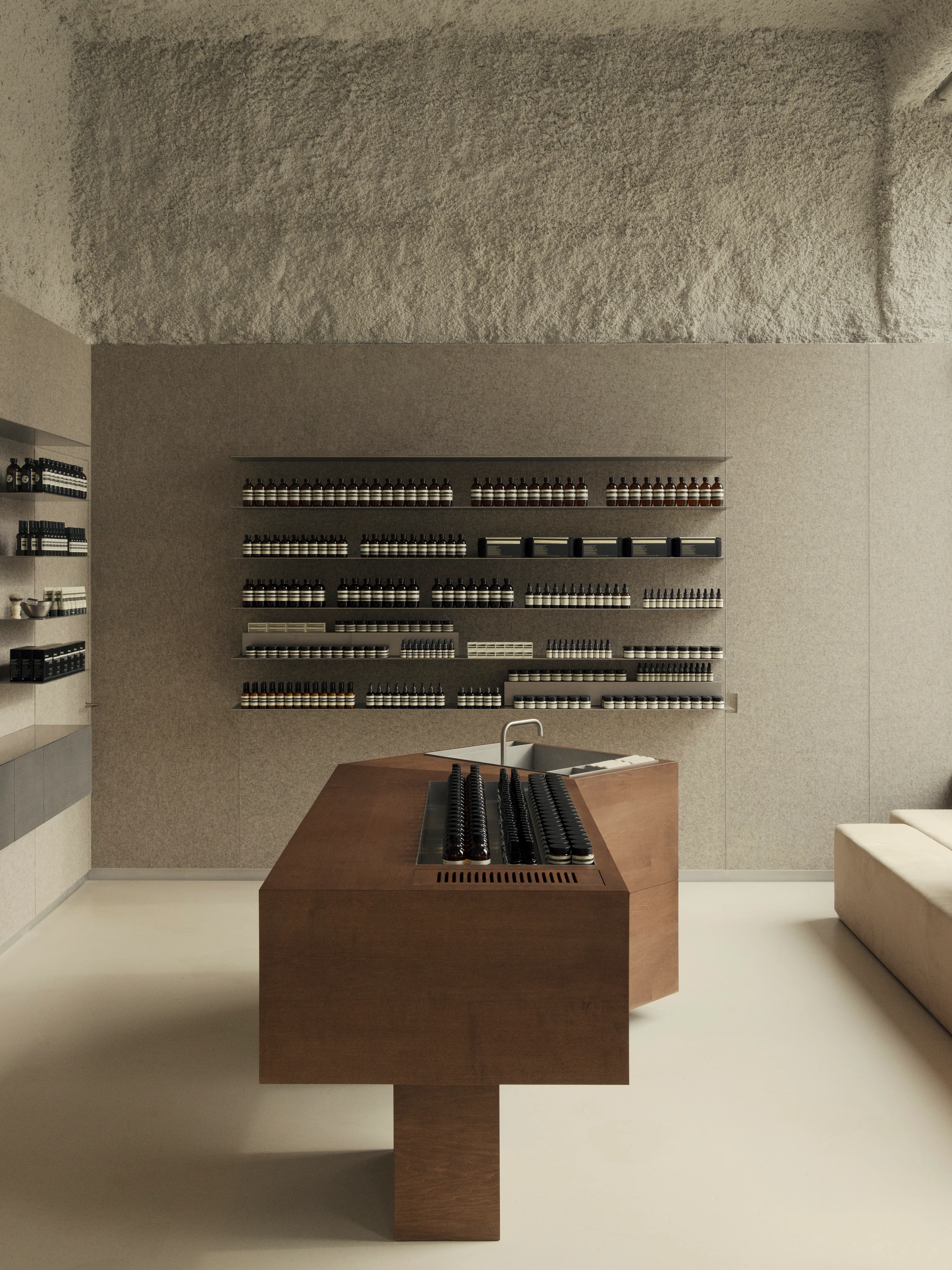 Minimalist retail interior with textured plaster ceiling, felt-paneled walls, and rows of amber glass apothecary bottles and skincare products displayed on floating shelves, centered around a dark wood island counter with a built-in sink, and a cream bench seating area on the right. Aesop Nicolas Schuybroek