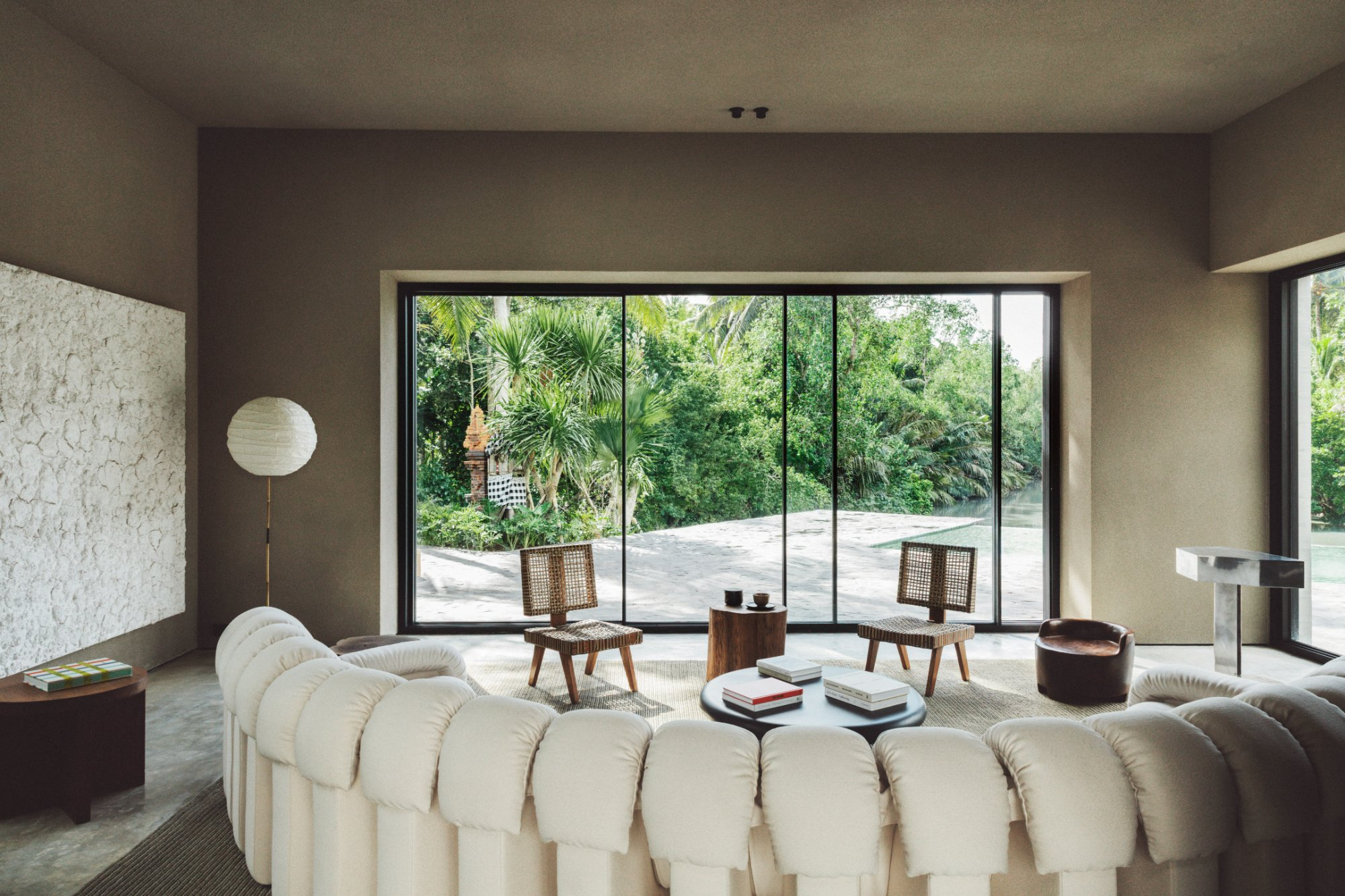 Modern living room with a large tufted cream sectional sofa, a round black coffee table with books, two woven rattan side chairs, a floor lamp with a round paper shade, and a floor-to-ceiling window framing a lush tropical garden with palm trees and a stone courtyard. Nicolas Schuybroek