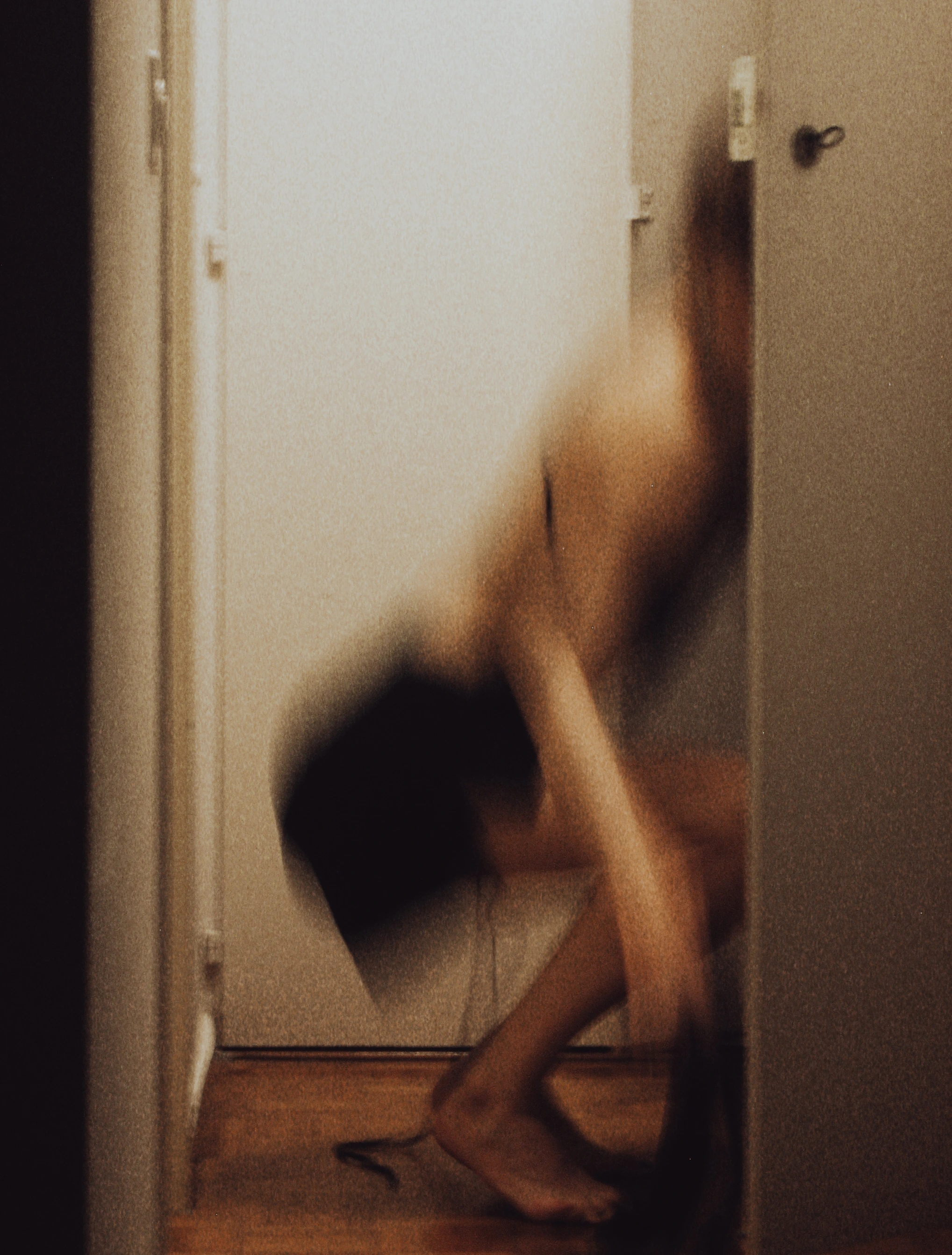 A blurred image of a barefoot figure in black shorts squats down in a hallway in front of an open white door, partially covered by another door. Serhii Tykhoniuk