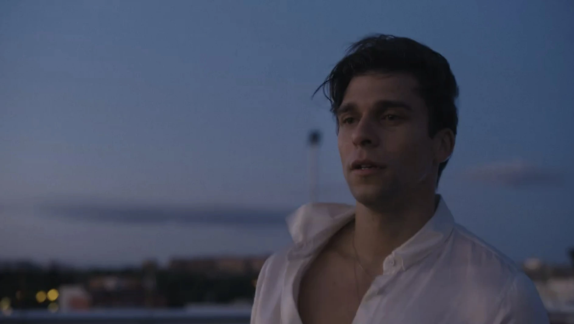 A man wearing an unbuttoned white shirt stares longingly ahead with his lips parted and a deep blue sky behind him. Valentino Sandoli