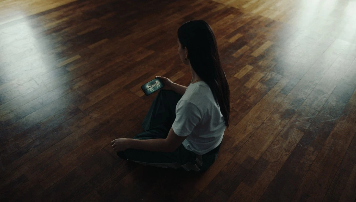 A woman sits cross-legged on a wooden floor, looking down at a phone that she is holding in her right hand. Jesse Plum
