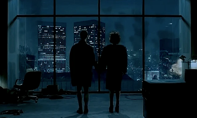 Silhouetted couple standing hand in hand before a wall of windows overlooking a nighttime city skyline, with buildings visibly exploding and collapsing in the background, from the final scene of