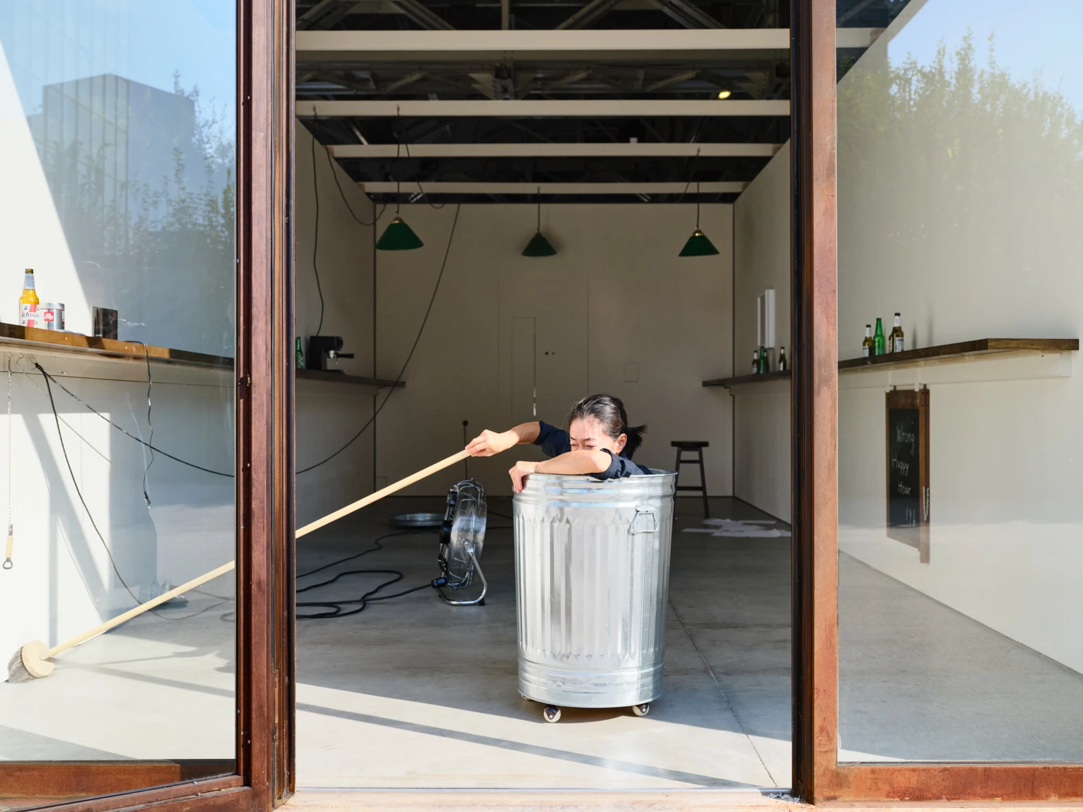 Aki Sasamoto performs Wrong Happy Hour, leaning over a metal trash can with a wooden stick in a Milan gallery installation.