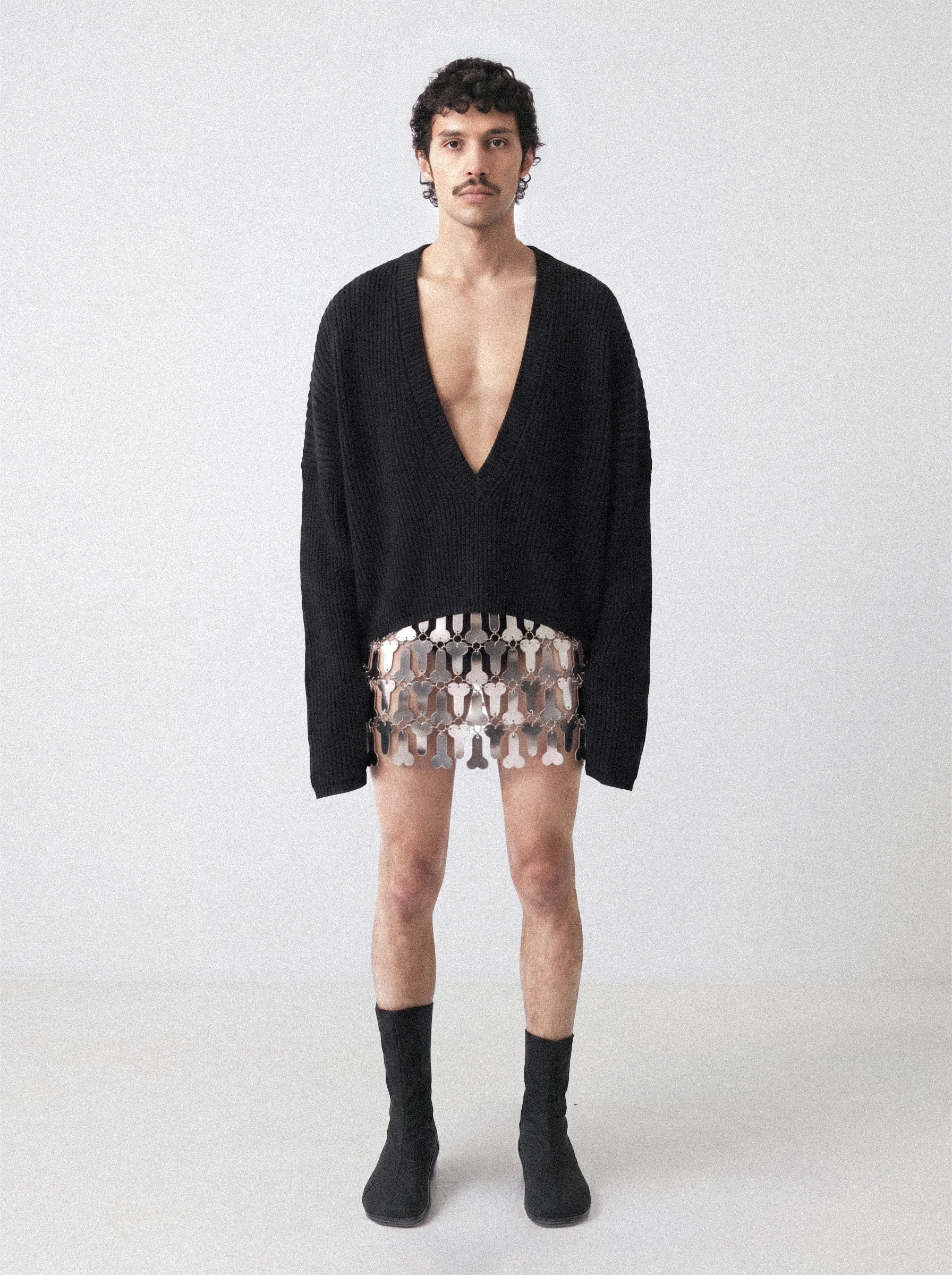 Goksenin Ay wearing a deep V-neck black knit sweater and silver chainmail mini skirt from debut collection Lament
