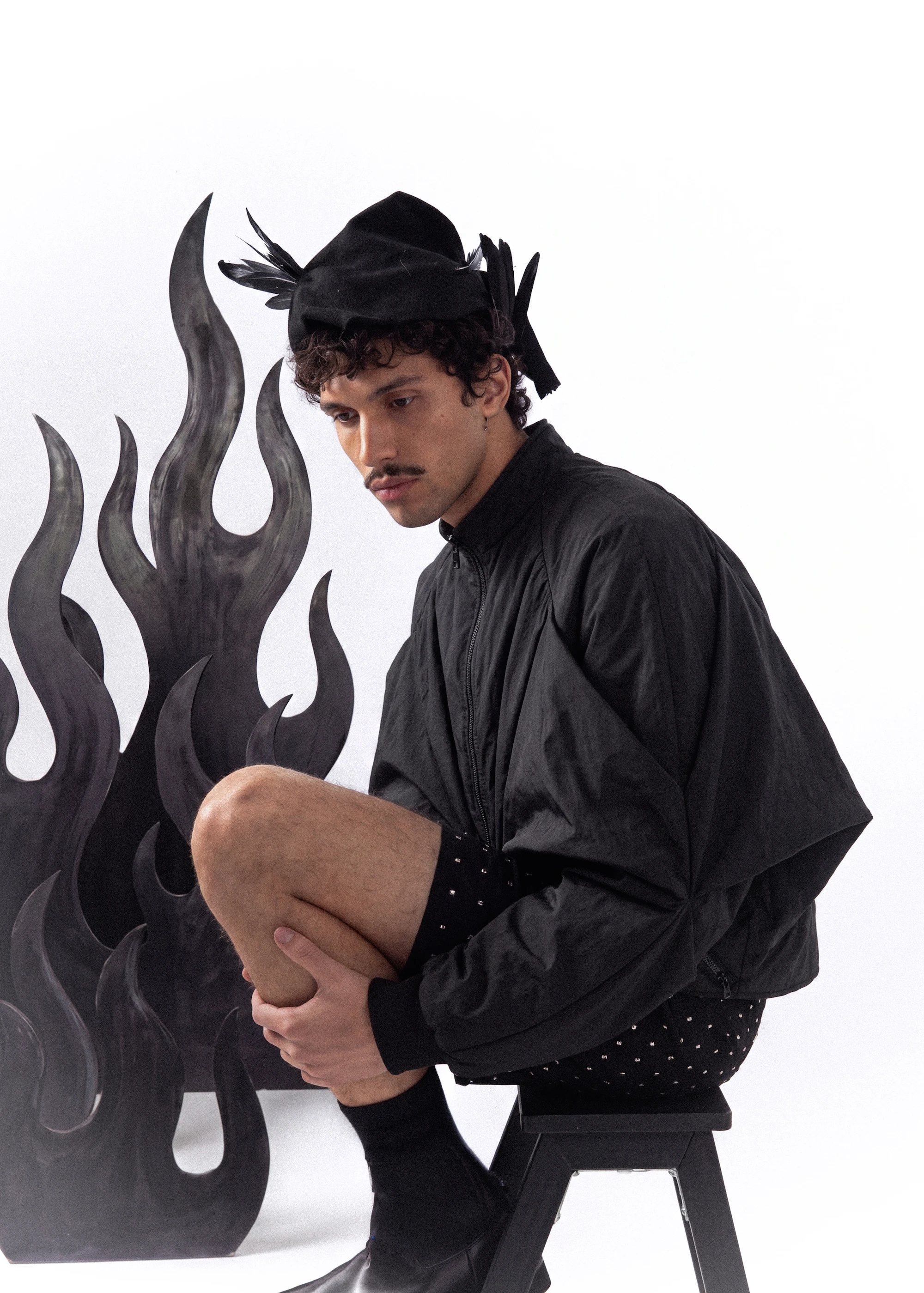 Goksenin Ay seated in black bomber jacket and feathered cap against dark flame backdrop, Lament debut collection