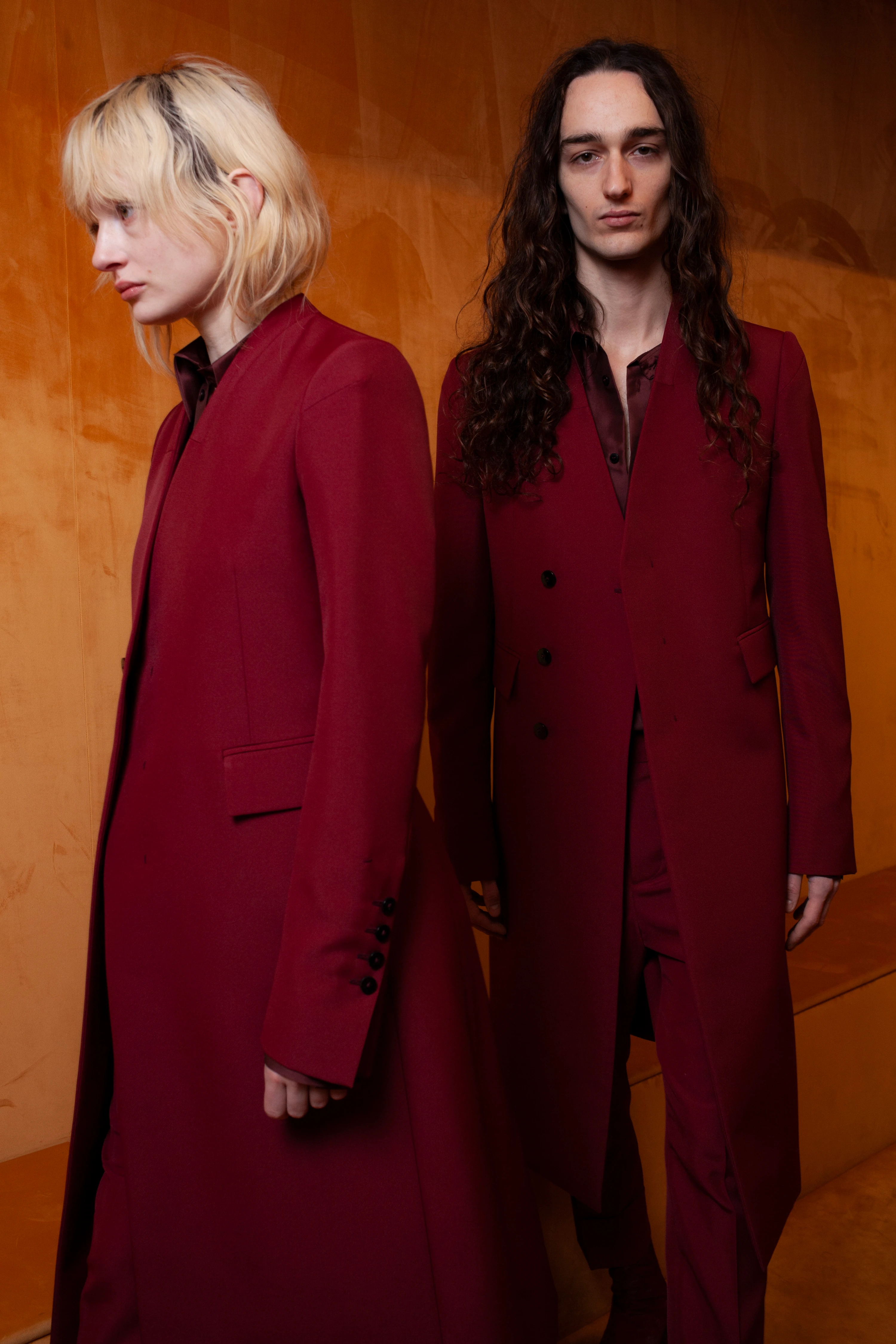 Two models in deep crimson tailored coats, SAPIO FW24 collection, against warm amber backdrop