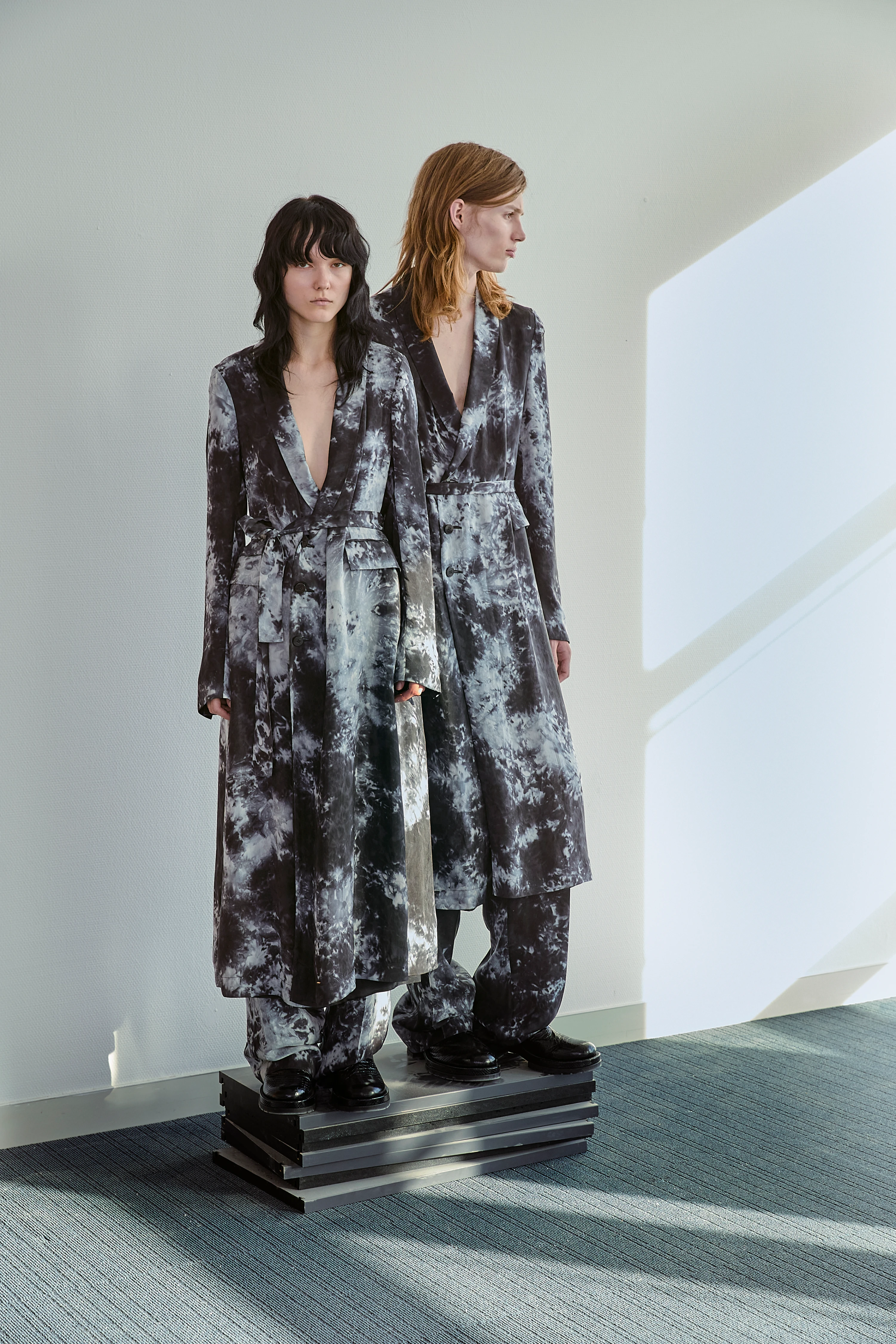 Two models wearing SAPIO SS25 matching dark tie-dye long coats and wide-leg trousers standing on a platform