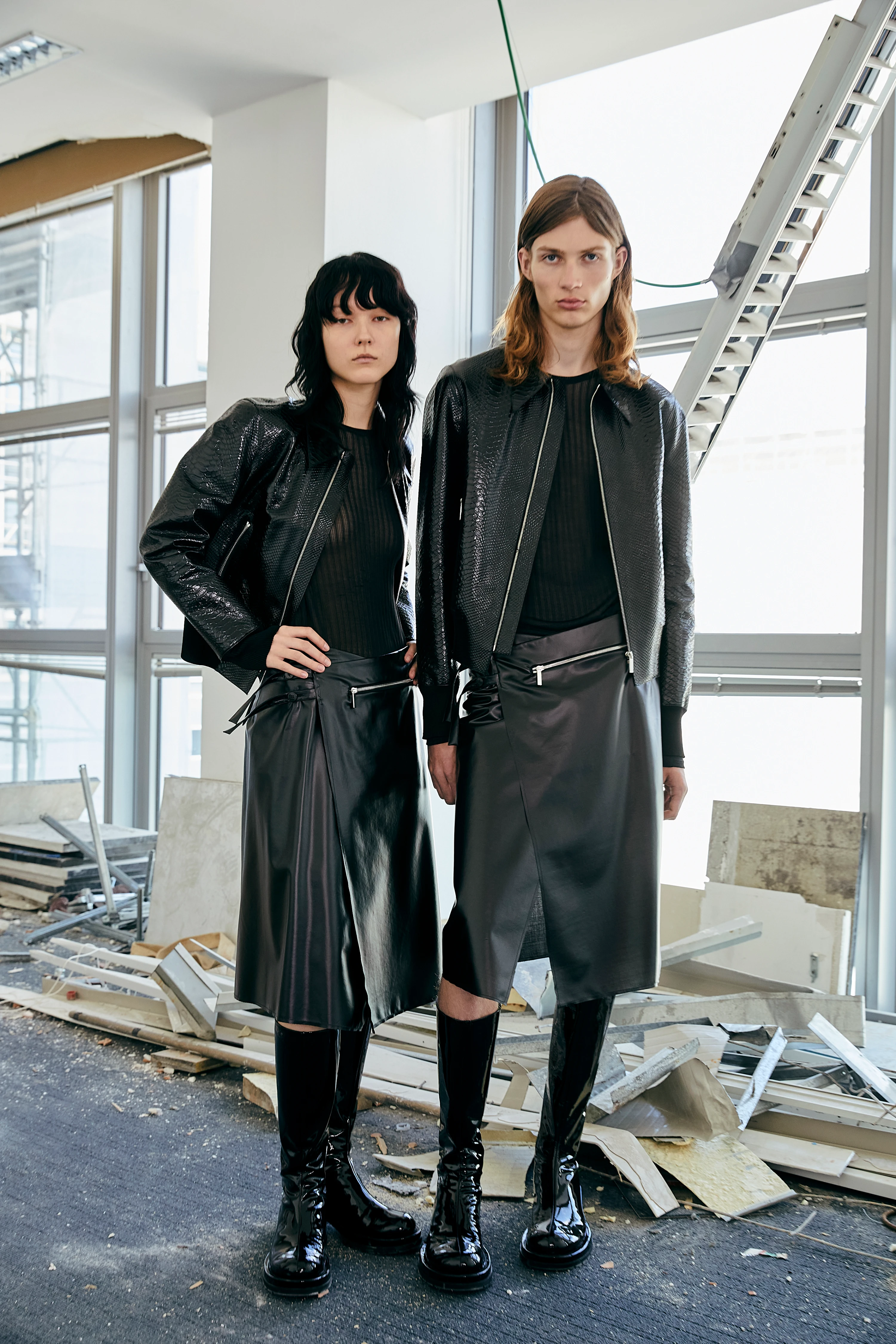 Two models wear SAPIO SS25 all-black leather midi skirts, sequin bombers, and patent boots in an industrial space.