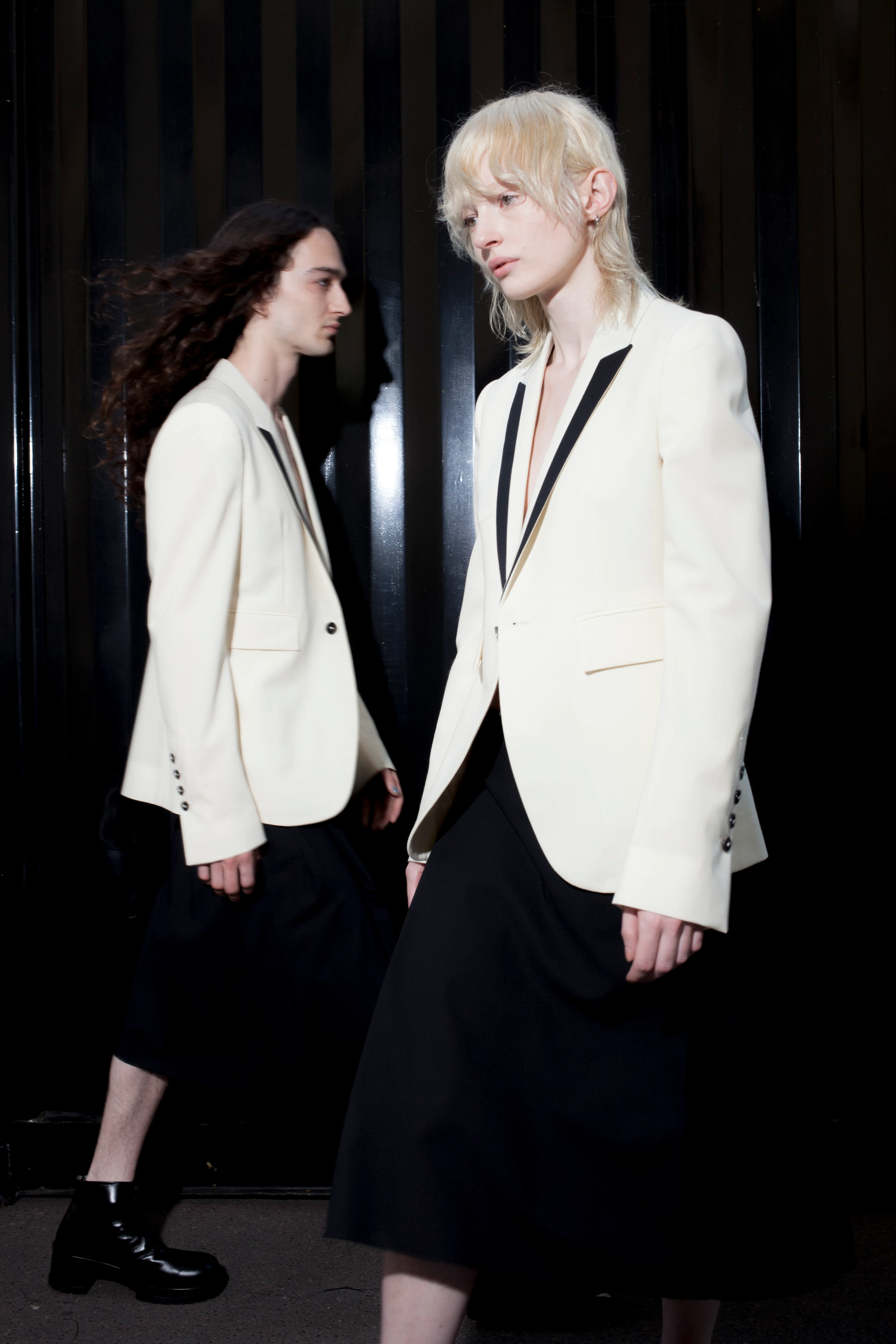 Two models wearing SAPIO SS24 ivory blazers with black lapels and dark skirts against a dramatic dark backdrop