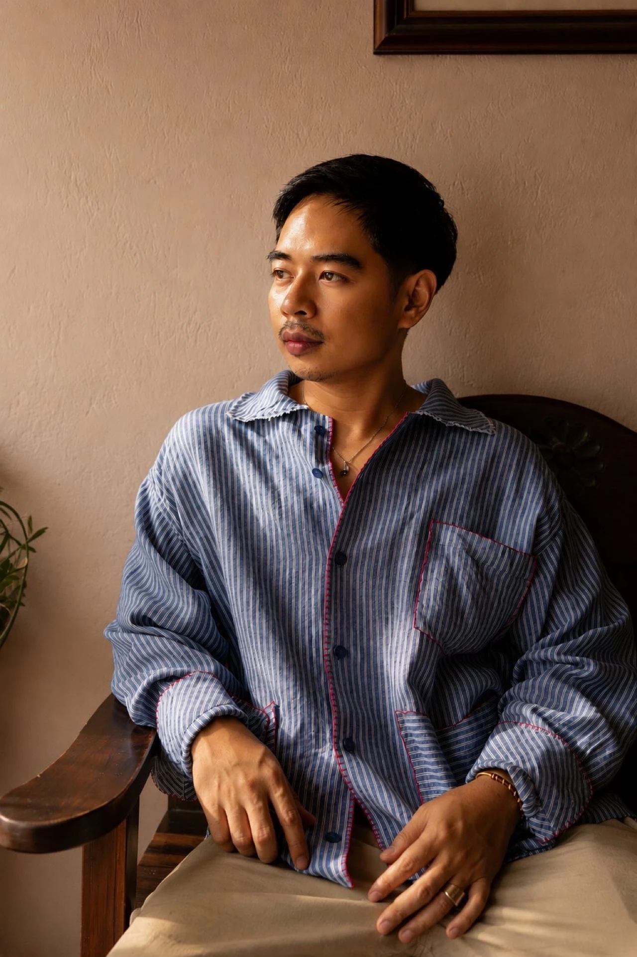Joseph Bagasao seated in wooden chair wearing blue striped overshirt, contemplative expression, warm ambient light