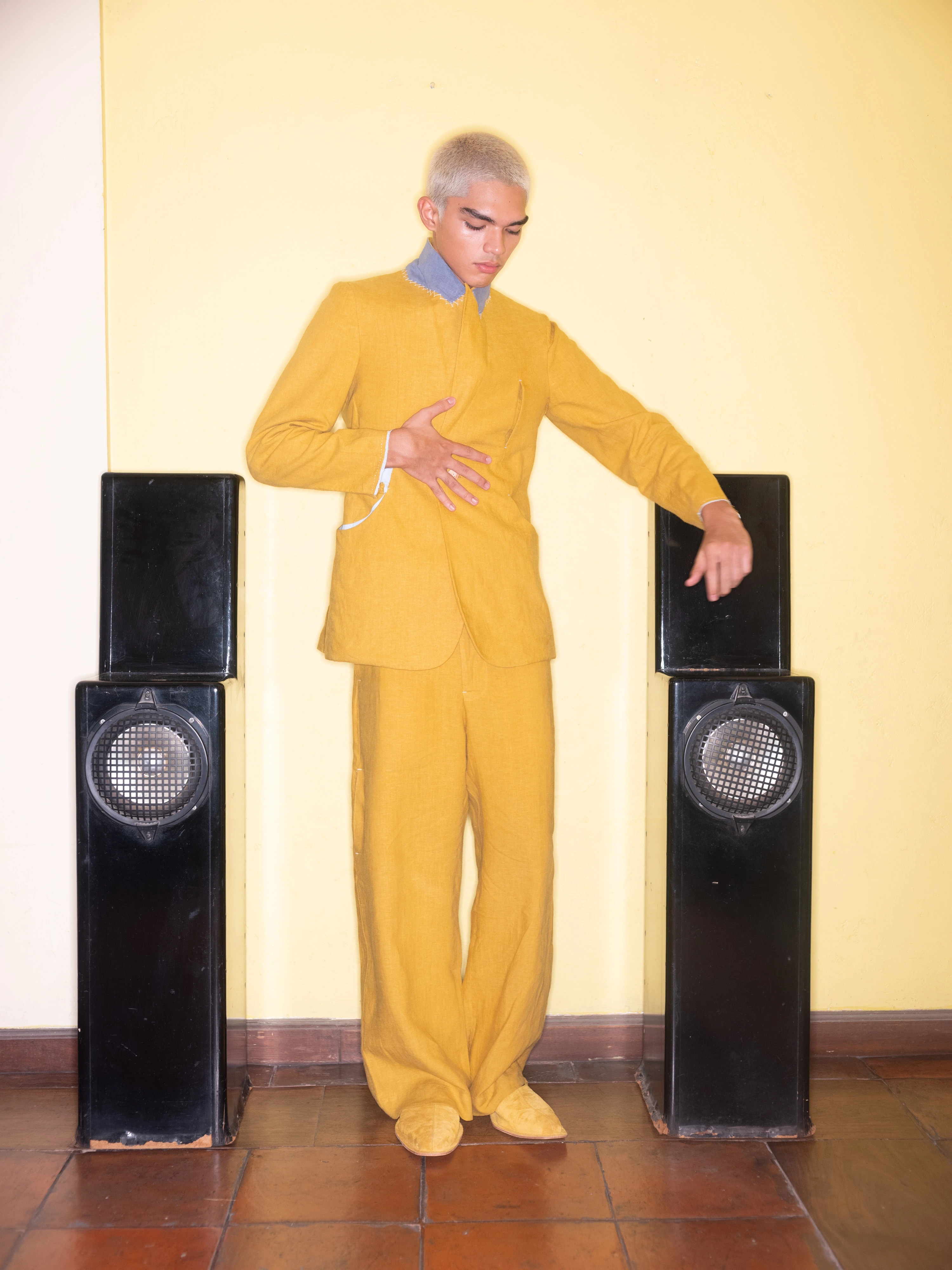 BAGASAO model in mustard yellow suit with bleached hair posed between vintage black tower speakers