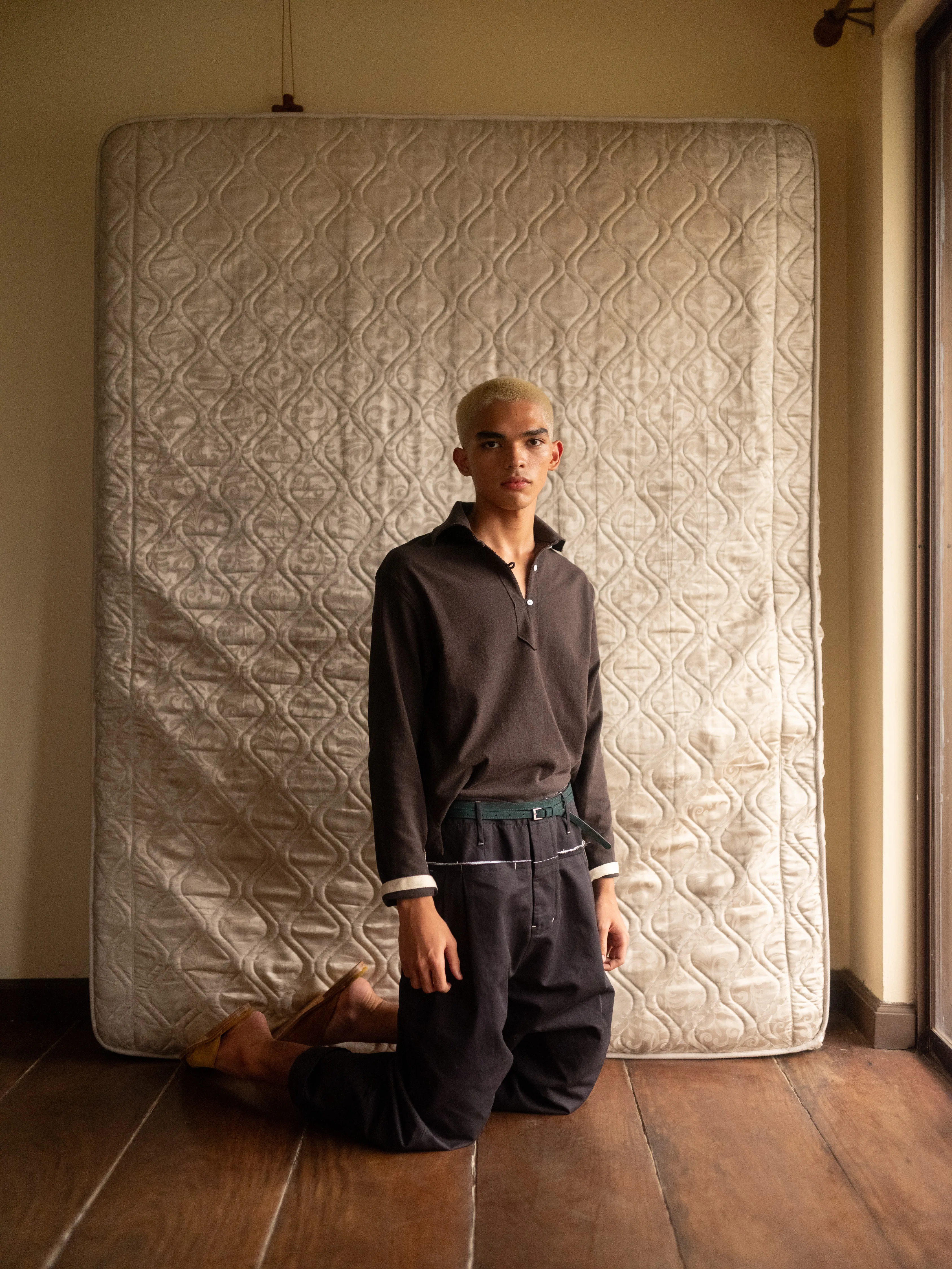 BAGASAO menswear model kneeling in dark polo shirt and wide-leg trousers against a quilted mattress backdrop