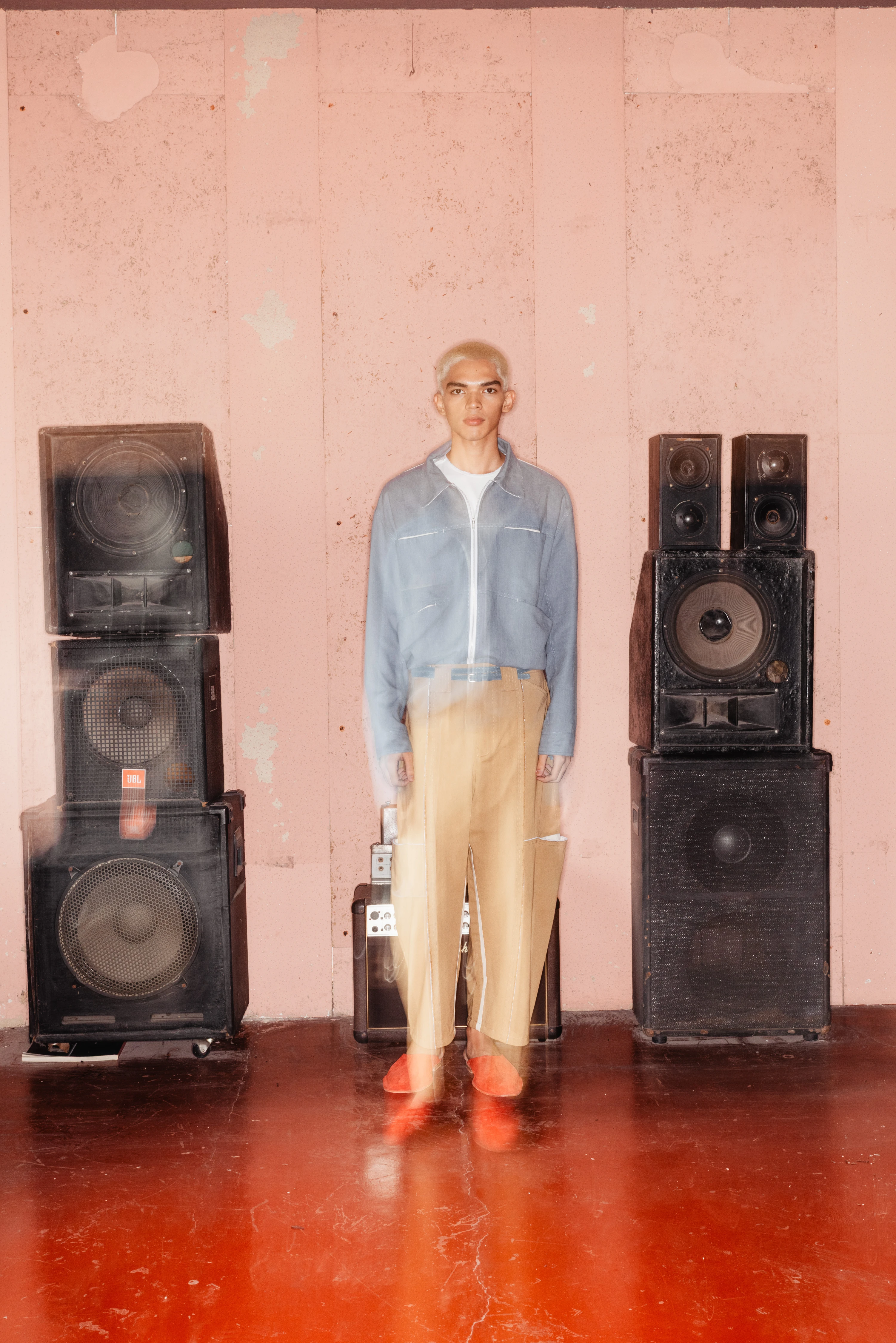 BAGASAO model in light blue jacket and tan trousers standing between vintage speaker stacks on red floor